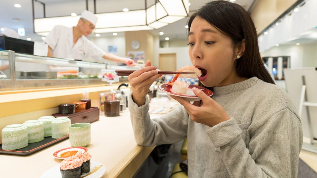A woman eating conveyor belt sushi and a sushi chef in the background
